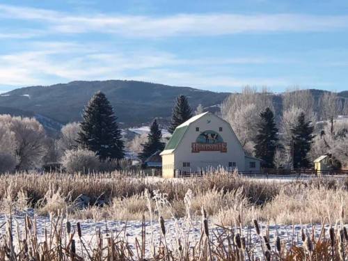 A picturesque barn with a green roof, surrounded by frosty trees and mountains under a clear blue sky.