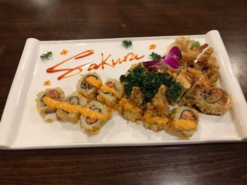 A beautifully arranged sushi platter featuring various rolls, tempura, and garnishes, with "Sakura" written in sauce.