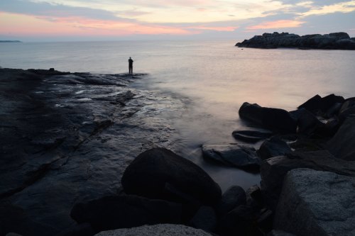 A lone figure stands on rocky shore at sunset, with calm waters and a colorful sky in the background.