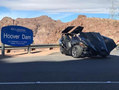 A sleek black vehicle parked near the Hoover Dam sign, with rocky terrain and blue sky in the background.
