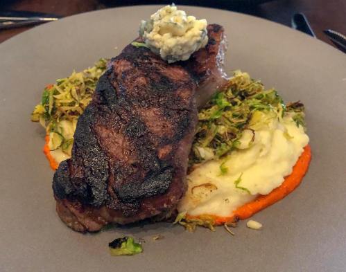 A grilled steak topped with blue cheese, served on a bed of mashed potatoes and sautéed Brussels sprouts.