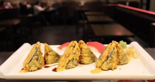 A plate of crispy sushi rolls drizzled with sauce, garnished with wasabi and pickled ginger, in a restaurant setting.