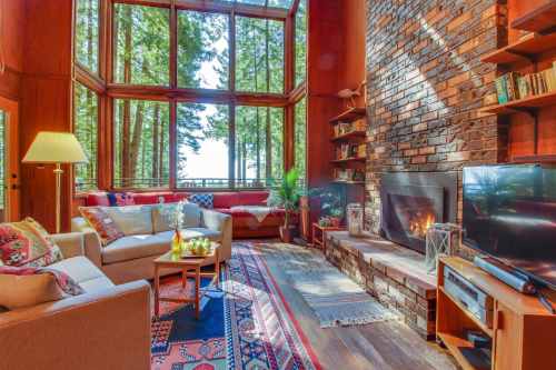 Cozy living room with large windows, a fireplace, and wooden accents, surrounded by trees and natural light.