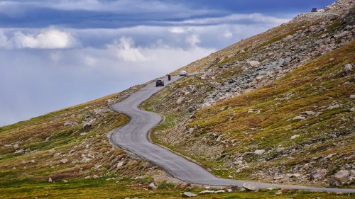 A winding road on a mountain slope, surrounded by rocky terrain and patches of grass under a cloudy sky.