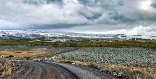 A winding dirt road leads through a landscape of rolling hills, patches of snow, and overcast skies.