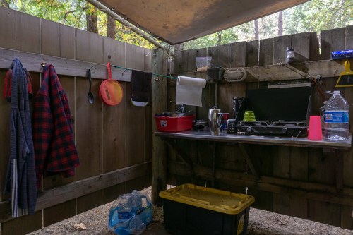 A rustic outdoor kitchen setup with a wooden table, cooking supplies, and hanging clothes in a forested area.