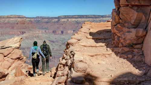 Two hikers explore a rocky trail at the Grand Canyon, with vast canyon views in the background under a clear blue sky.