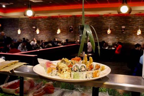 A platter of assorted sushi sits on a counter, with a busy restaurant scene in the background.