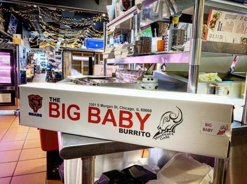 A large box labeled "The Big Baby Burrito" sits on a counter in a restaurant, with festive lights in the background.