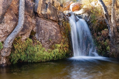 A serene waterfall cascades over rocks into a tranquil pool, surrounded by lush greenery and rugged terrain.