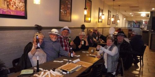A group of people celebrating at a restaurant, wearing party hats and holding drinks, smiling at the camera.