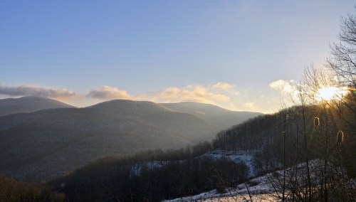 A serene mountain landscape at sunrise, with rolling hills and a clear blue sky, partially covered in snow.