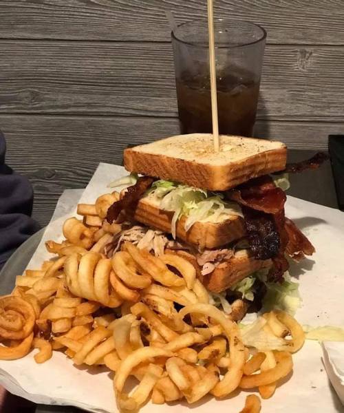 A towering sandwich with layers of lettuce, bacon, and pulled meat, served with a generous portion of curly fries.