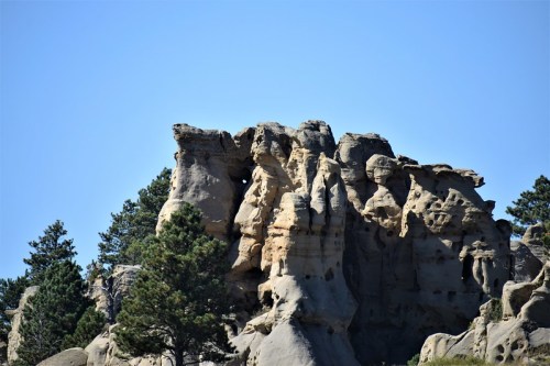 Rocky cliff formation with trees in the foreground against a clear blue sky.