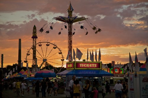 A vibrant fair scene at sunset, featuring rides, ticket booths, and colorful tents with people enjoying the festivities.