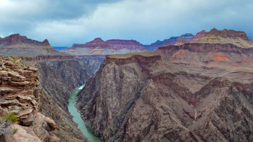 A dramatic view of a canyon with steep cliffs and a winding river below, under a cloudy sky.