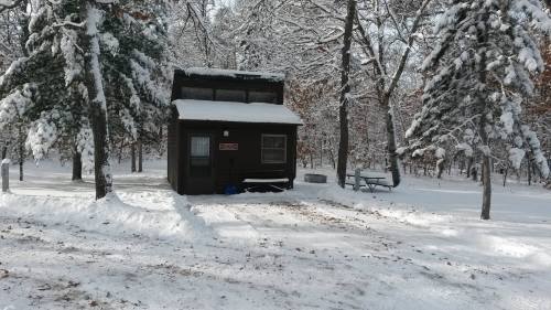 A small cabin surrounded by snow-covered trees in a winter landscape. Snow blankets the ground and roof.