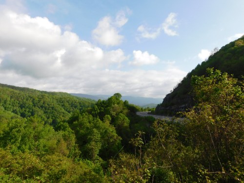 A scenic view of lush green mountains under a partly cloudy sky, with a winding road visible in the distance.