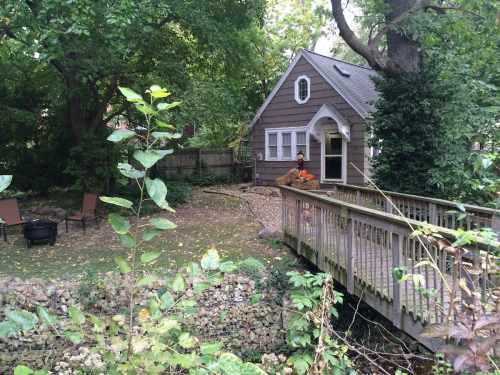 A cozy house surrounded by greenery, with a wooden bridge leading to a small patio area.