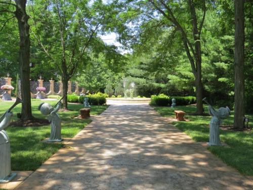 A serene garden path lined with statues and trees, leading to a fountain in the distance.
