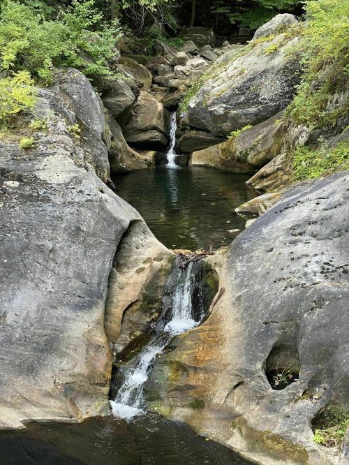 A serene waterfall flows between large rocks, creating a tranquil pool surrounded by lush greenery.