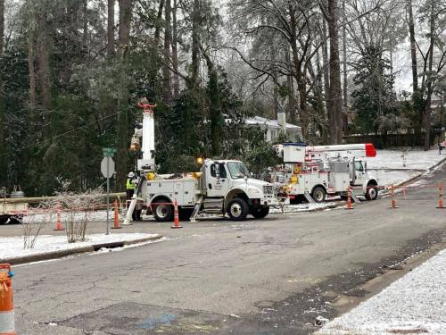 Two utility trucks with cherry pickers on a snowy street, surrounded by traffic cones and trees.