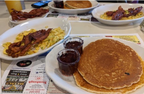 A table with plates of pancakes, scrambled eggs, bacon, and syrup in small cups.