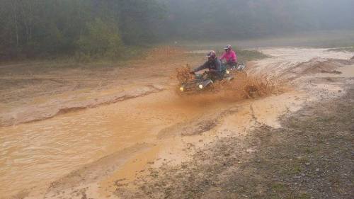 Two riders on an ATV splash through a muddy puddle in a foggy outdoor setting.