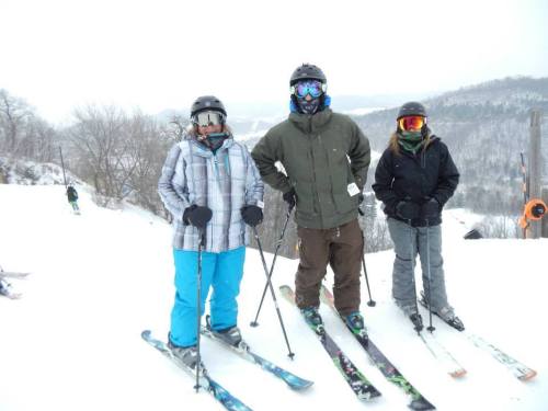 Three skiers pose on a snowy slope, wearing winter gear and goggles, with a snowy landscape in the background.