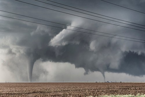 Two tornadoes touch down in a stormy sky over a flat landscape, with power lines in the foreground.