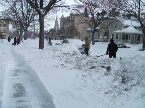 People shoveling snow from a sidewalk and street, surrounded by deep snowdrifts and bare trees in winter.