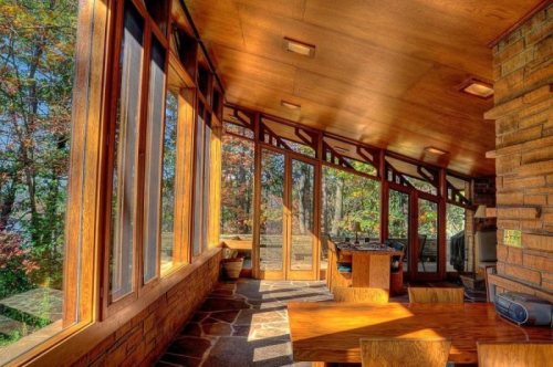 Bright, wooden sunroom with large windows overlooking a forest, featuring a stone floor and wooden furniture.