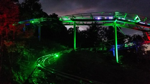 A brightly lit roller coaster track winds through trees at night, illuminated by red and green lights.