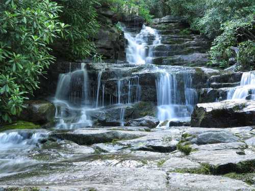 A serene waterfall cascades over rocky steps, surrounded by lush greenery and moss-covered stones.