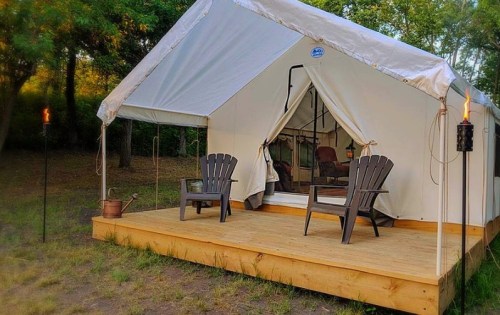 A cozy tent on a wooden deck, with two chairs and tiki torches, surrounded by trees in a serene outdoor setting.