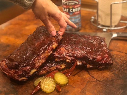 A hand reaches for a stack of glazed ribs on a wooden cutting board, accompanied by pickles and peppers.