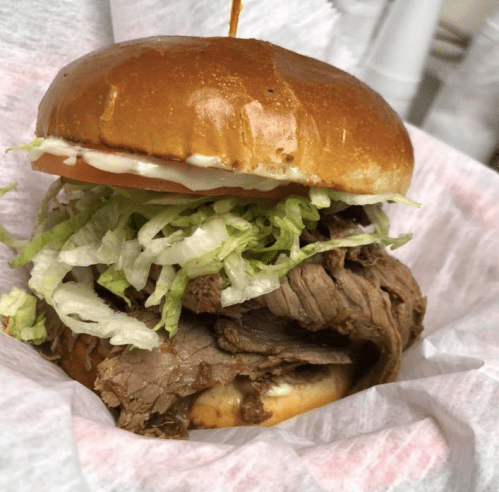 A close-up of a beef sandwich with lettuce and tomato on a toasted bun, served in a paper wrapper.