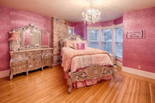 A glamorous bedroom with pink glitter walls, a silver bed, and a vintage dresser, featuring a chandelier and large windows.