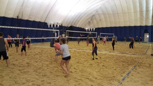 A busy indoor sand volleyball court with players engaged in a game under a large dome structure.