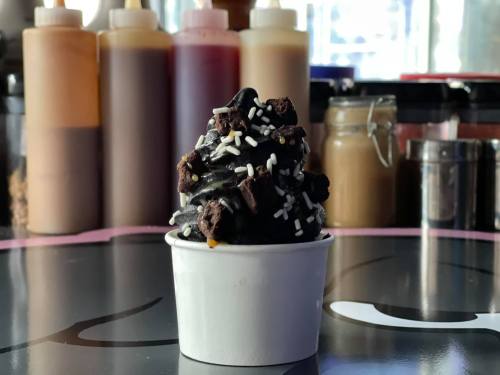 A small cup of dark chocolate ice cream topped with chocolate chunks and colorful sprinkles, set against a blurred background.