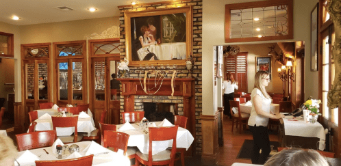 A cozy restaurant interior with wooden tables, a fireplace, and a large painting on the wall. A server is attending to guests.