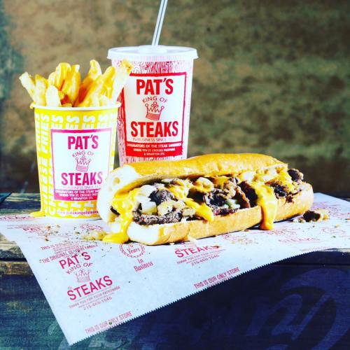 A cheesesteak sandwich with melted cheese, fries, and a drink from Pat's King of Steaks on a branded paper.