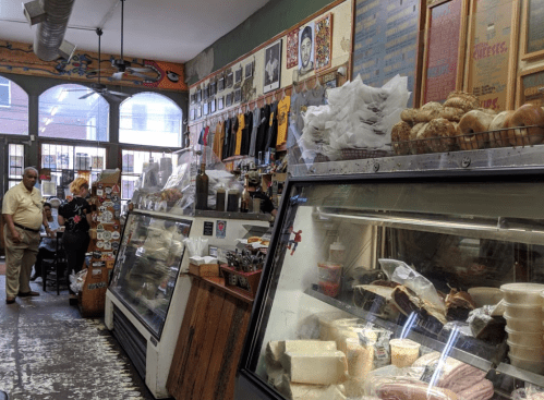 A deli interior with glass displays of meats and cheeses, colorful decor, and customers browsing in a lively atmosphere.