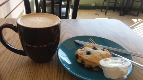 A coffee mug beside a blue plate with a blueberry scone and a small container of cream on a wooden table.