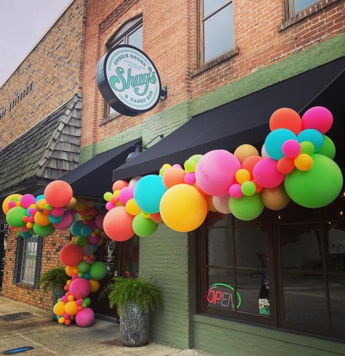 Colorful balloon arch outside a candy shop named "Shrugs," with an "OPEN" sign in the window.