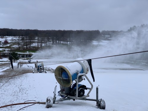 Snowmaking machines operate on a snowy slope, creating artificial snow against a cloudy sky.
