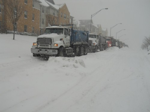 A line of trucks stuck in deep snow during a heavy snowstorm, with residential buildings in the background.