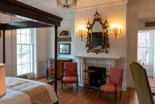A cozy bedroom featuring a four-poster bed, two pink chairs, a fireplace, and elegant decor with natural light.