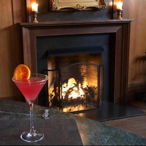 A pink cocktail with an orange slice sits on a table in front of a cozy fireplace. Candles glow softly in the background.
