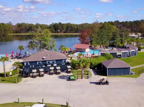 Aerial view of a lakeside resort with buildings, a pool, and golf carts near palm trees and colorful umbrellas.